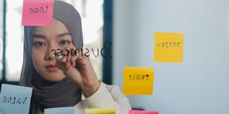 Asia muslim lady write information, strategy, reminder on glass board in new normal office. Working from home, remotely work, self isolation, social distancing, quarantine for coronavirus prevention.